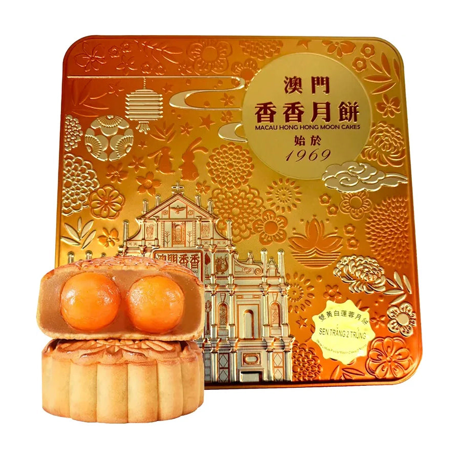 澳门香香双黄白莲蓉月饼礼盒，金色铁盒包装印有大三巴牌坊图案，前方展示切开的月饼，露出两颗饱满的咸蛋黄。
