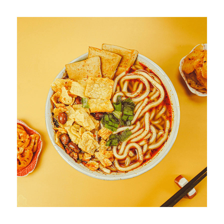 俯拍视角下的砂锅土豆粉成品特写，白色碗中盛有红油汤底、粗土豆粉、豆皮、花生和葱花，旁边配有筷子和小菜。