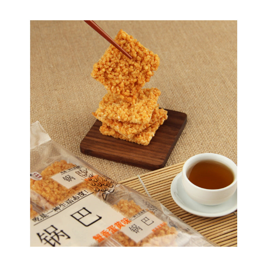 筷子夹起一块金黄酥脆的方形锅巴特写，背景为木质托盘、部分产品包装袋及一杯清茶。