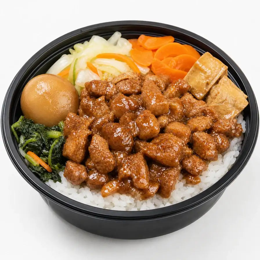 俯拍视角下的火车便当，黑色圆碗内盛满白米饭，上方铺有红烧肉丁、一颗卤蛋、两块卤豆腐、炒青菜、胡萝卜片和卷心菜。